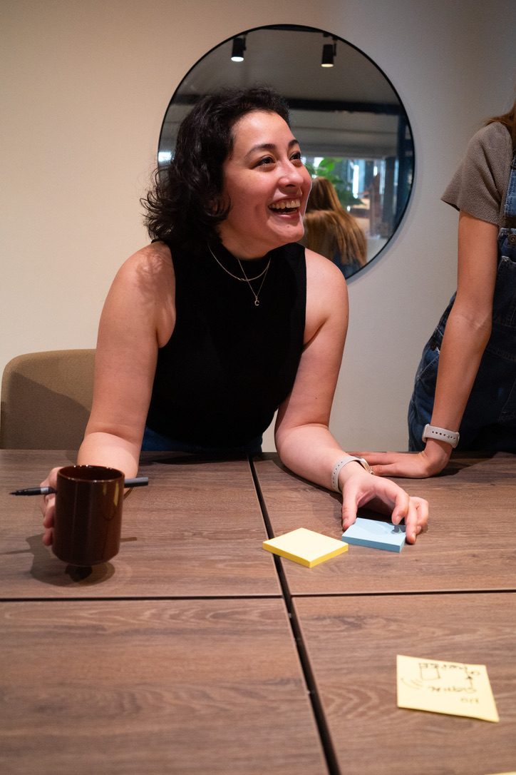 Cristina laughing with a workshop group, leaning on a table covered in sticky notes